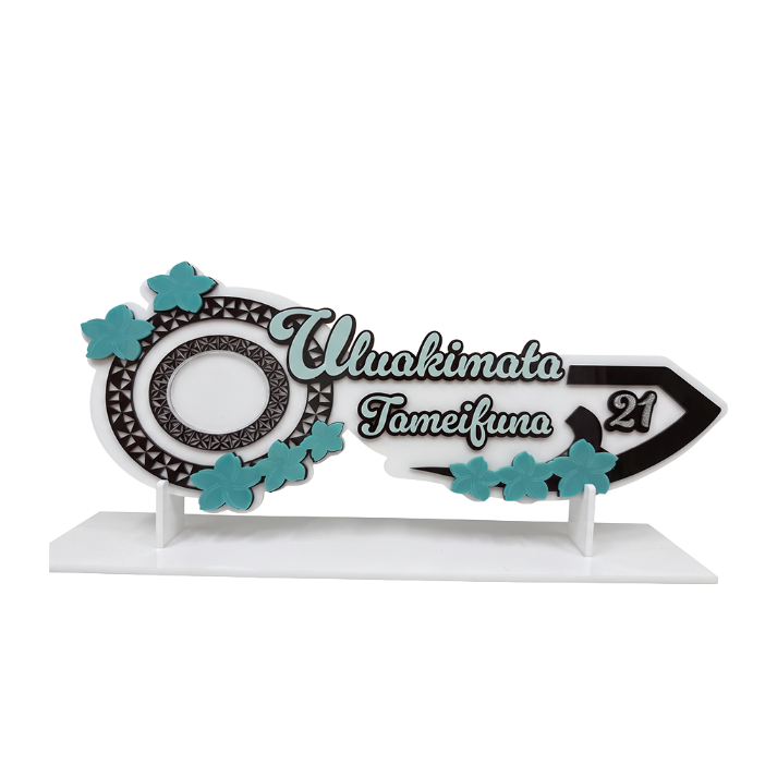 A decorative custom sign or plaque on a white display stand, featuring the text 'Uluakimata Jameifuna 21' in white and black script over a black and white patterned circular and arrowhead design, accented with teal butterfly shapes, on a white background.
